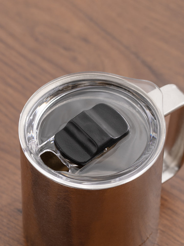 Close-up of a stainless steel travel mug with a clear lid and black flip-top spout, resting on a wooden surface.