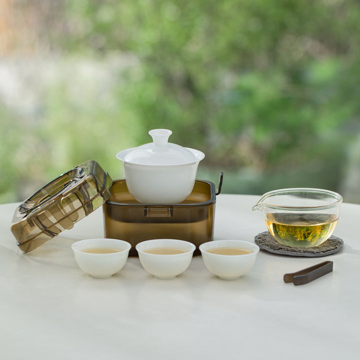 A tea set arrangement on a white table with a white lidded teapot, three small white cups, a clear glass pitcher filled with tea, a dark metal container, and a bamboo lid, against a blurred green outdoor background.