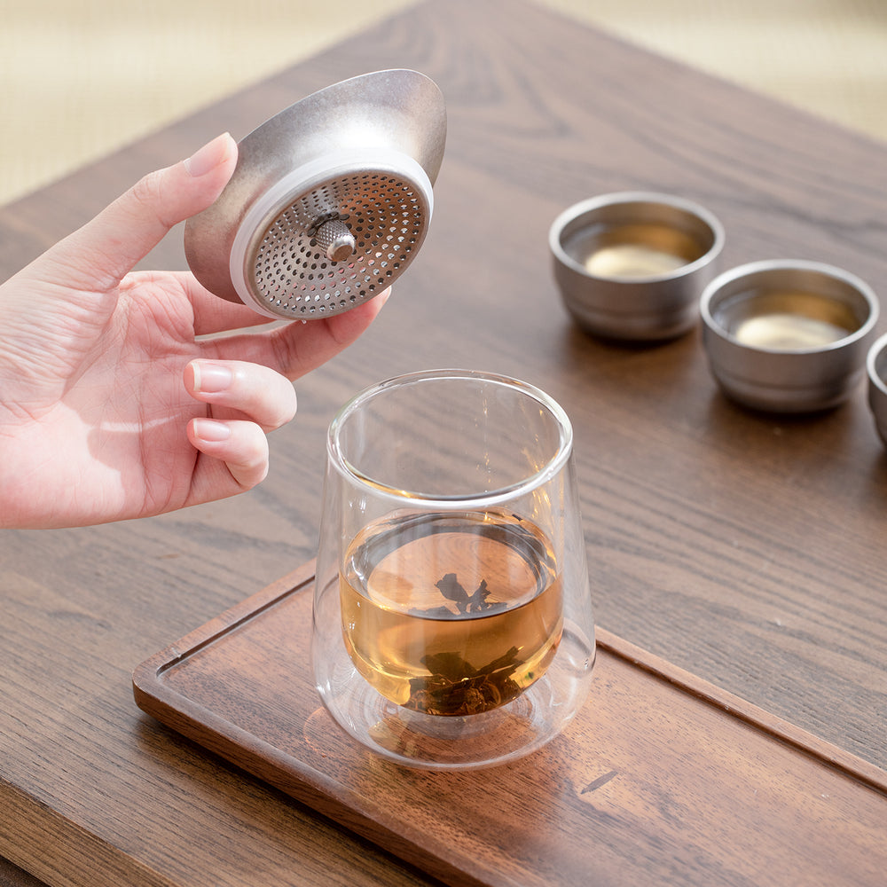 Person holding a tea infuser over a glass of tea on a wooden surface.