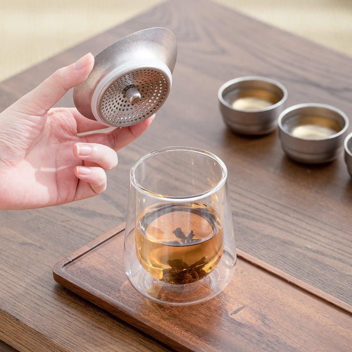 Person holding a tea infuser over a glass of tea on a wooden surface.