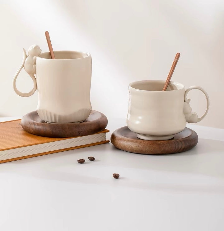 Two white cat mugs with wooden coasters on a light surface, accompanied by coffee beans and a book.
