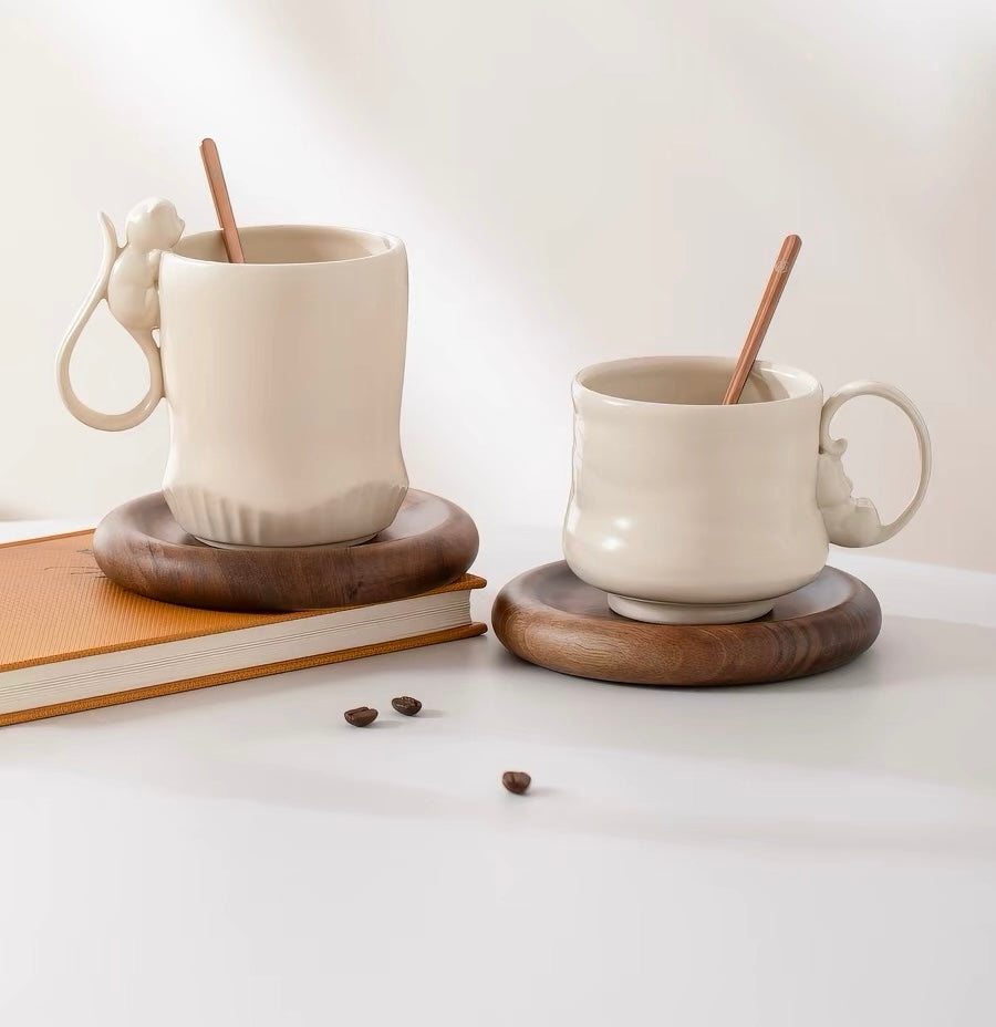 Two white cat mugs with wooden coasters on a light surface, accompanied by coffee beans and a book.