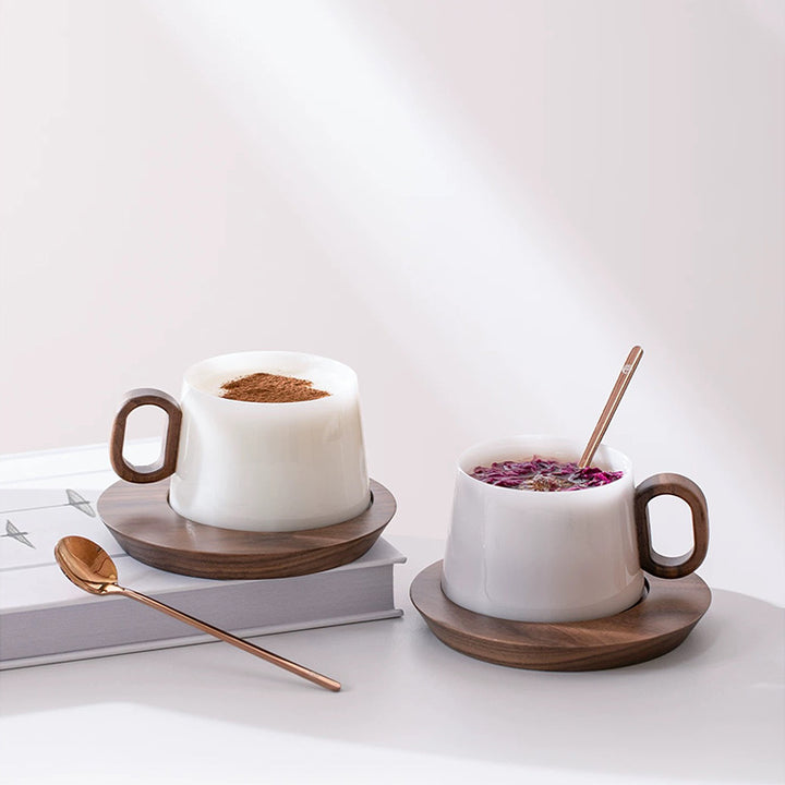 Two white mugs with wooden handles on saucers, filled with a dessert and a floral arrangement, on a light surface.