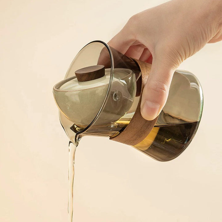 Glass teapot with wooden handle and lid being poured from a hand on a beige background