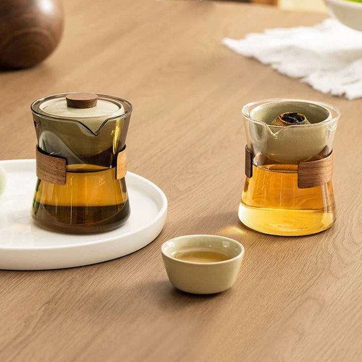 Tea-making set with glass teapot, wooden handle, and small white cup on a wooden surface.