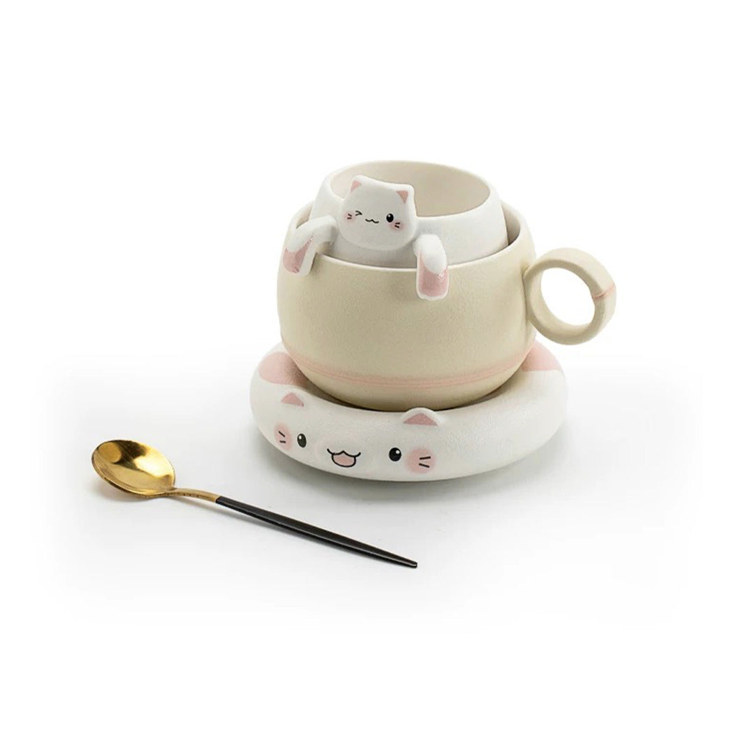 A beige ceramic cat-shaped mug with a white cat face design, accompanied by a ceramic saucer and a metal spoon, against a white background.