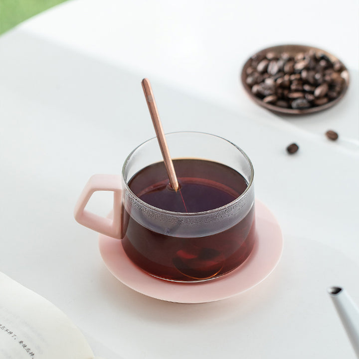 Glass cup of black tea with a pink saucer on a white surface, accompanied by a spoon and a small bowl of coffee beans.