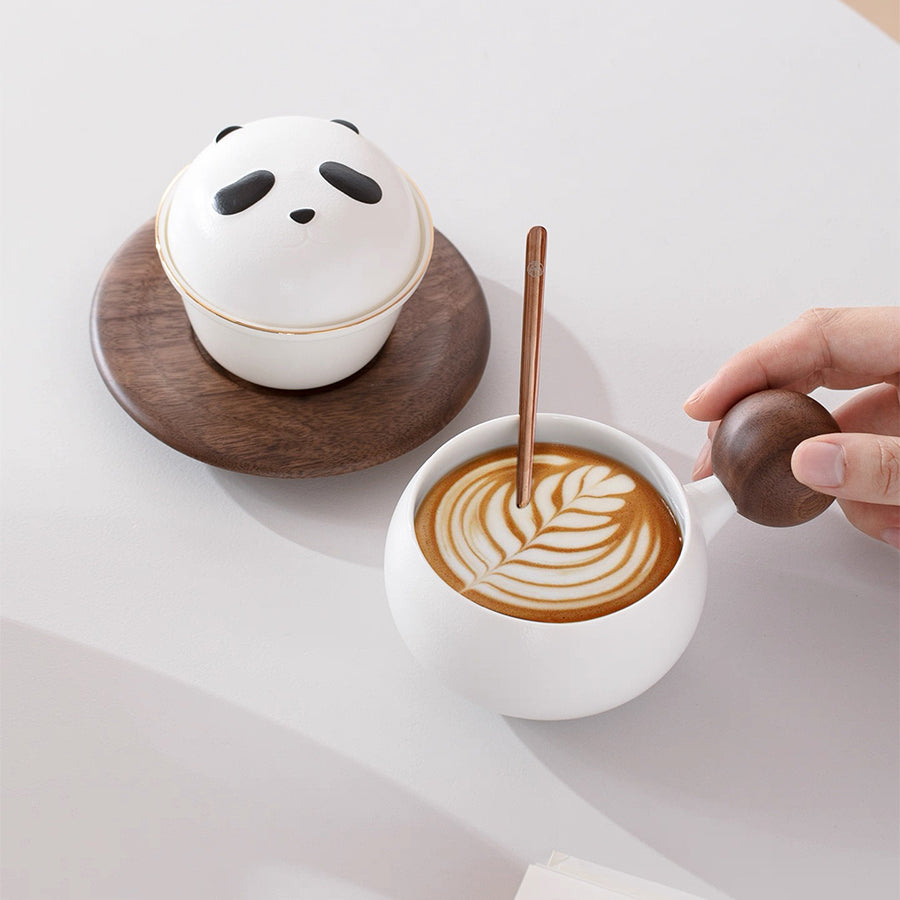 White panda-shaped coffee mug on a wooden coaster next to a cup of coffee with latte art.
