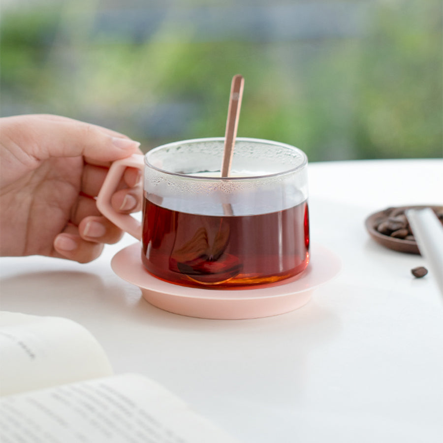 Glass mug with a pink base filled with tea, held by a hand on a blurred natural background.
