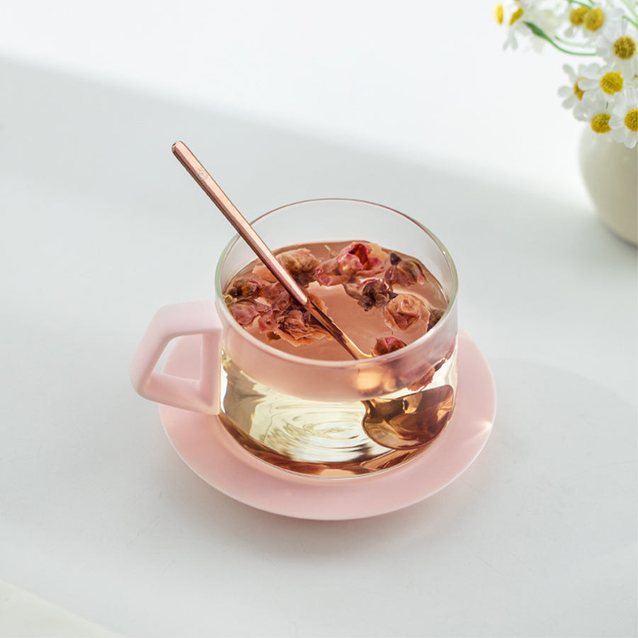 Clear glass teacup with pink handle and saucer containing a hot beverage with visible herbs, on a light gray background.