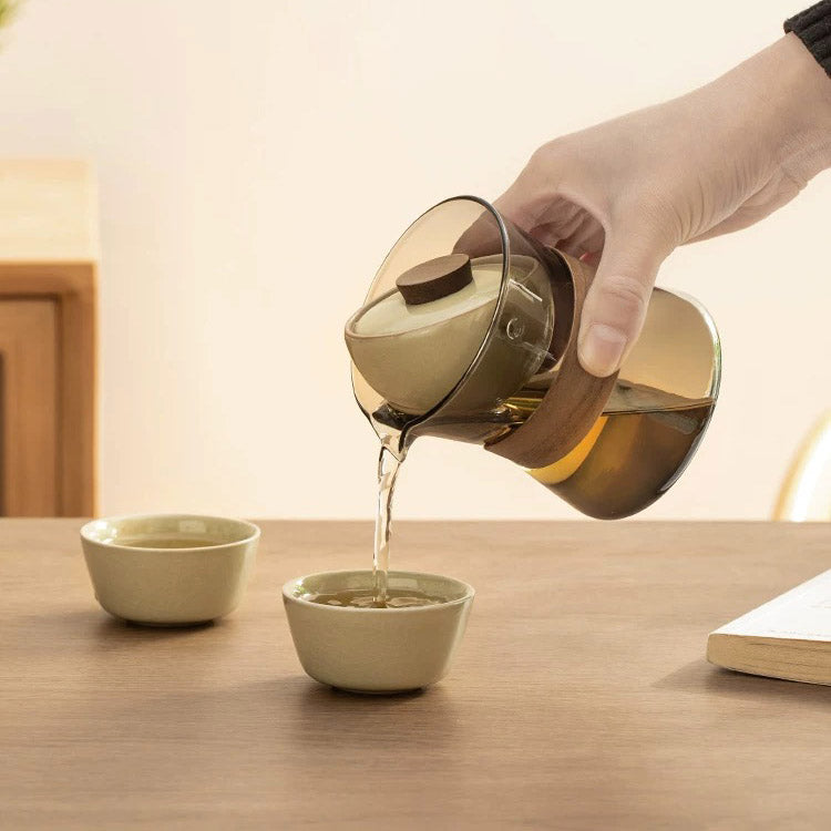 Person pouring tea from a glass teapot with rotate infuser into two ceramic cups on a wooden table.