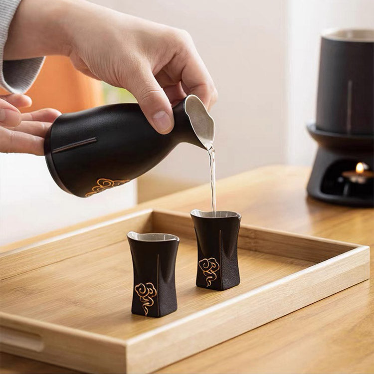 A person's hand pours liquid from a black ceramic sake pitcher with gold calligraphic details into two matching black and silver sake cups on a wooden tray.