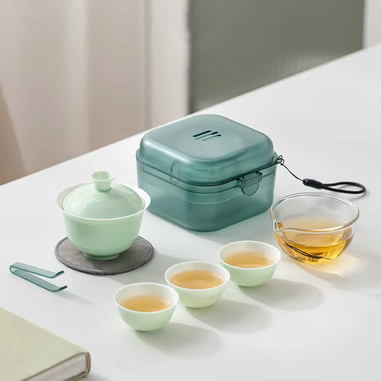 A tea set arranged on a white tabletop, including a pale green lidded teapot, a translucent mint-green storage box, a clear glass cup of amber tea, two small pale green tea cups, and a metal tea scoop. The scene is softly lit with a neutral indoor background.