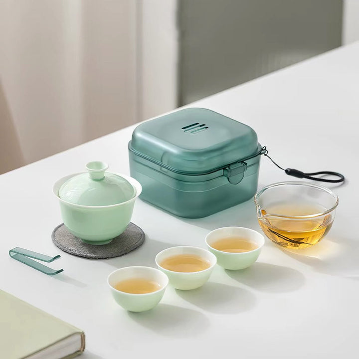 A tea set arranged on a white tabletop, including a pale green lidded teapot, a translucent mint-green storage box, a clear glass cup of amber tea, two small pale green tea cups, and a metal tea scoop. The scene is softly lit with a neutral indoor background.