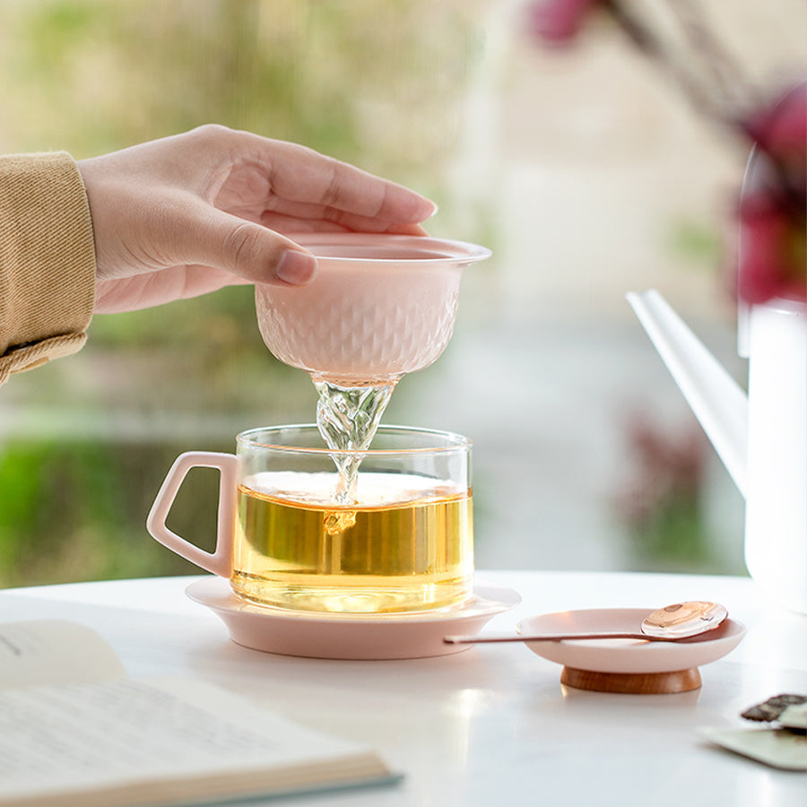 Person pouring tea from a pink teapot into a glass cup on a white table.
