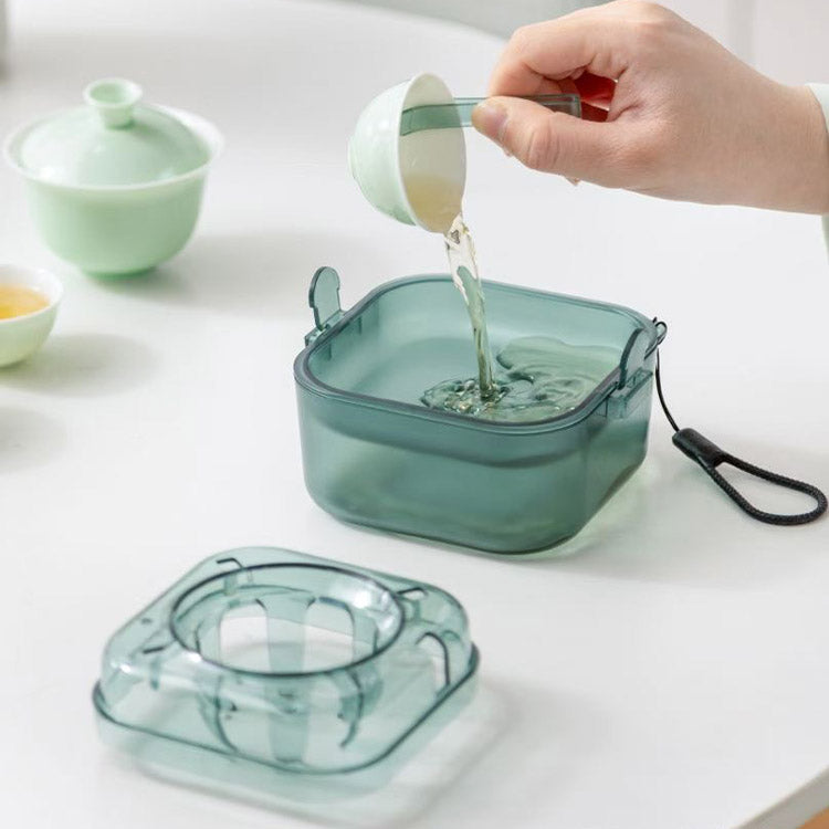 A translucent smoky green square tea storage container with a lid and carrying strap, shown on a white tabletop with a hand pouring tea into it. A matching lid and a light green teapot are also visible in the scene.