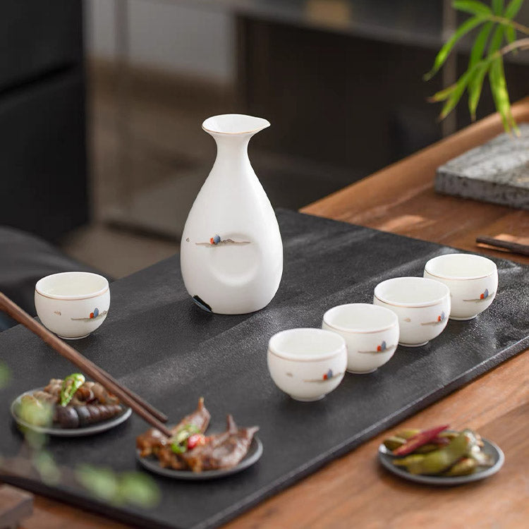 White ceramic sake set with a small bottle and five matching cups arranged on a black serving tray on a wooden table, with chopsticks and small dishes of food nearby.